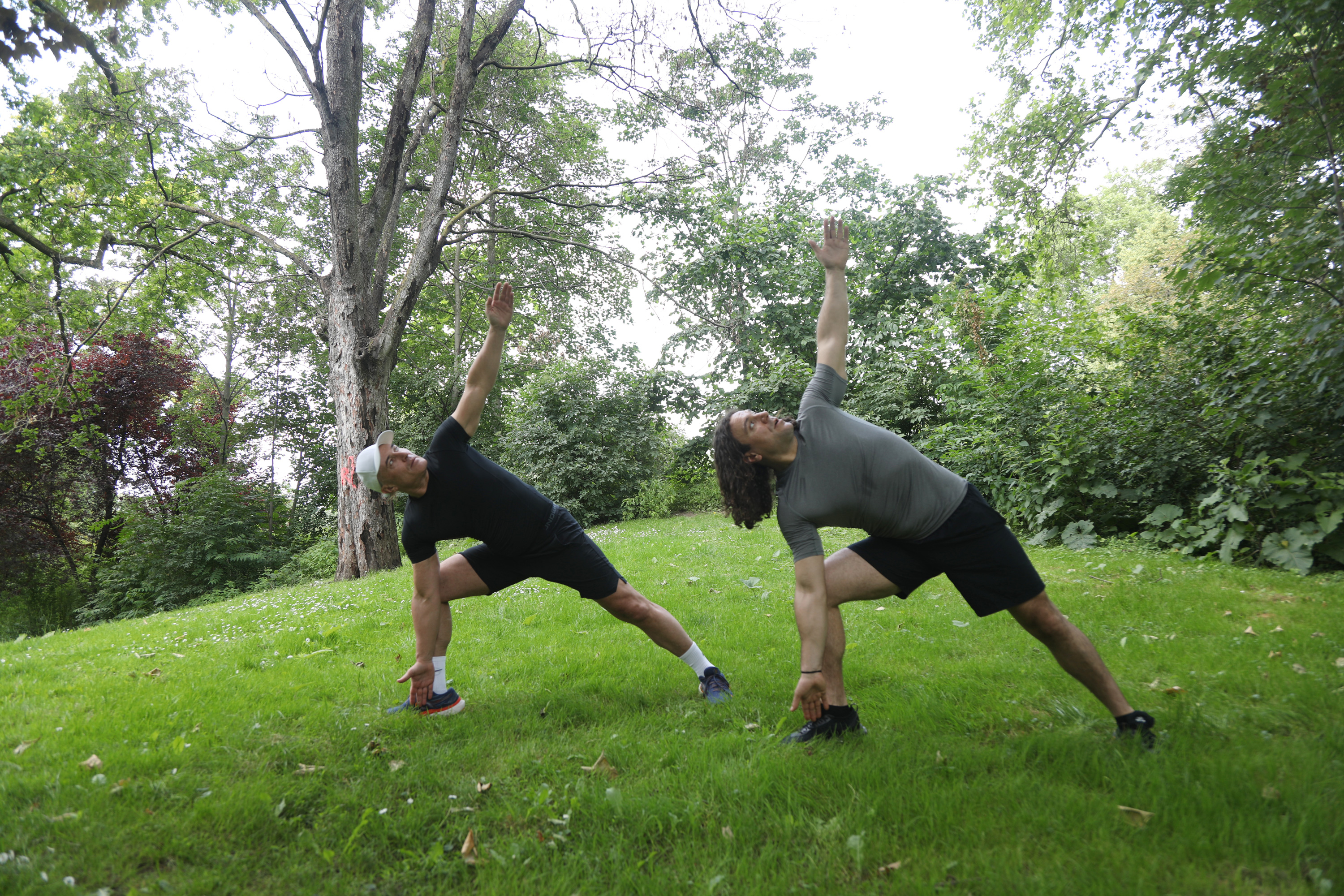Exercices de Yoga en Plein Air - Étirement Deux hommes pratiquent des exercices d'étirement en plein air sur une pelouse verte avec des arbres en arrière-plan.