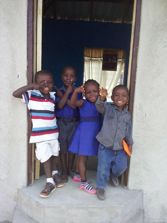 Enfants dans un Encadrement de Porte – 4 Personnes Quatre enfants heureux se tiennent devant une porte, souriant et posant. Les garçons portent des t-shirts rayés et unis, la fille est en robe bleue.