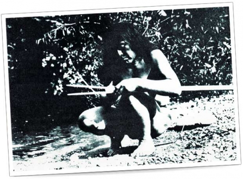 Méthode de chasse traditionnelle – Photo en noir et blanc Un homme est assis au bord d'une rivière en fabriquant une lance, pieds nus et vêtu de vêtements naturels.