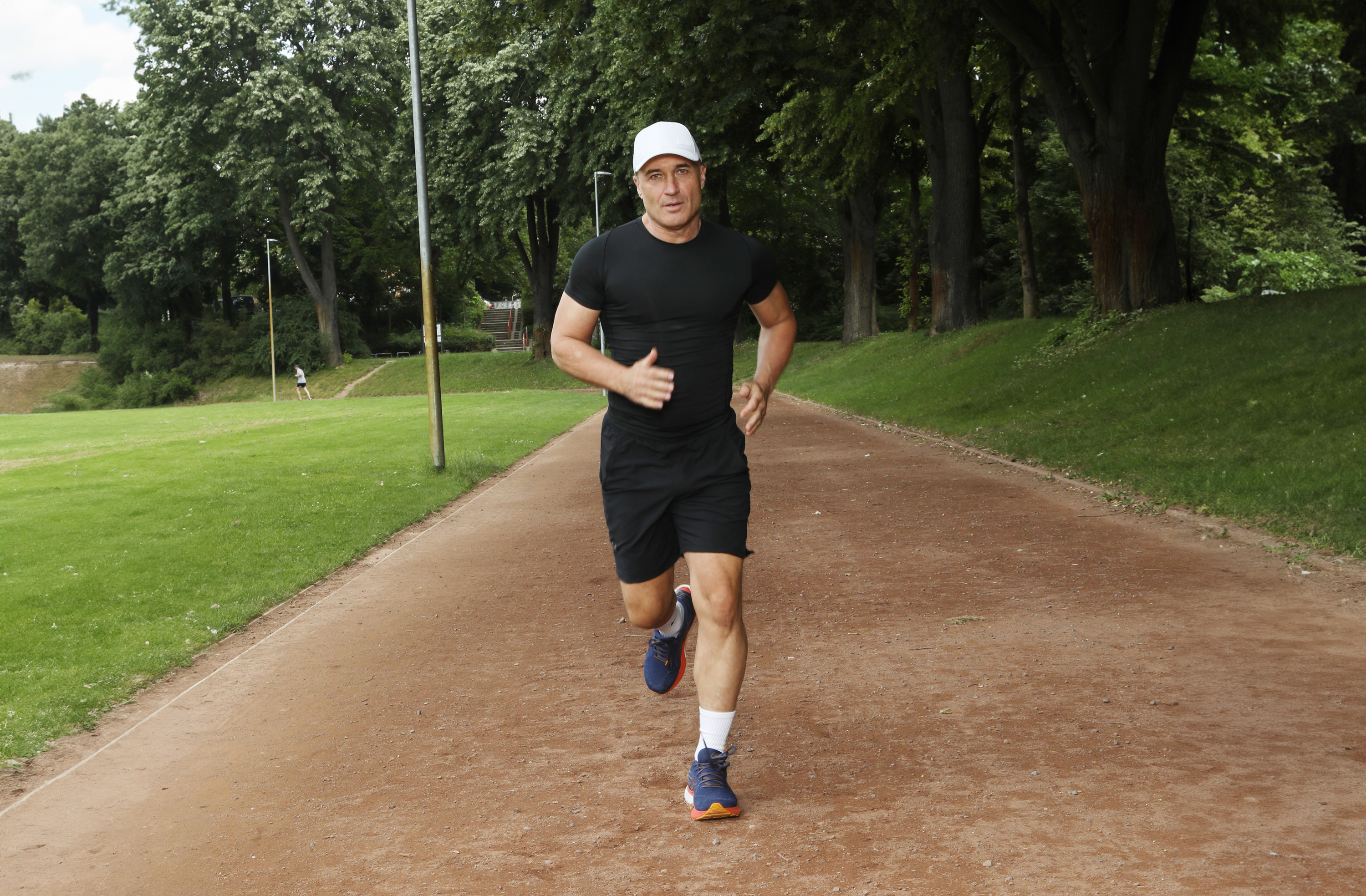 Chaussures de Course Hommes – Modèles Sportifs 10 km Homme courant sur un chemin forestier, portant des vêtements de sport noirs et des chaussures de course bleues.