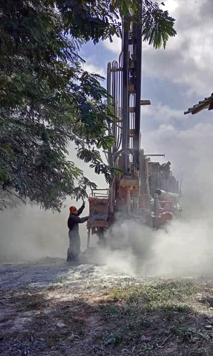 Perceuse – Forage d'eau sur site Un travailleur utilise une perceuse dans un environnement poussiéreux, avec des arbres en arrière-plan.